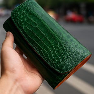 May include: A green leather wallet with a crocodile skin texture. The wallet is held in a hand, showcasing its detailed pattern and colour. The interior trim is a contrasting orange.