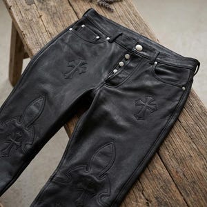 Peut inclure: Pantalon en cuir noir avec des ornements en forme de croix. Le pantalon est doté d'une braguette boutonnée et de poches. Des motifs de croix sont présents à l'avant et sur les jambes. Présenté sur une surface en bois.