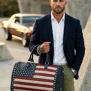 May include: A navy blue and red duffel bag with the American flag design. The bag features a leather exterior with silver-colored studs around the edges. It has black handles and a black shoulder strap. The bag is held by a person wearing a navy blue blazer and white shirt.