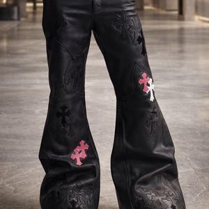 May include: Black leather flared trousers featuring multiple cross-shaped patches in black, white, and pink. The pants have a wide-leg silhouette with tonal stitching and decorative cross appliqués distributed across the front of both legs.