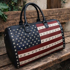 May include: A black, red, and white duffel bag with the American flag design. The bag features a navy blue section with gold stars, red and white stripes, and silver studs around the edges and on the flag design. It has black handles.