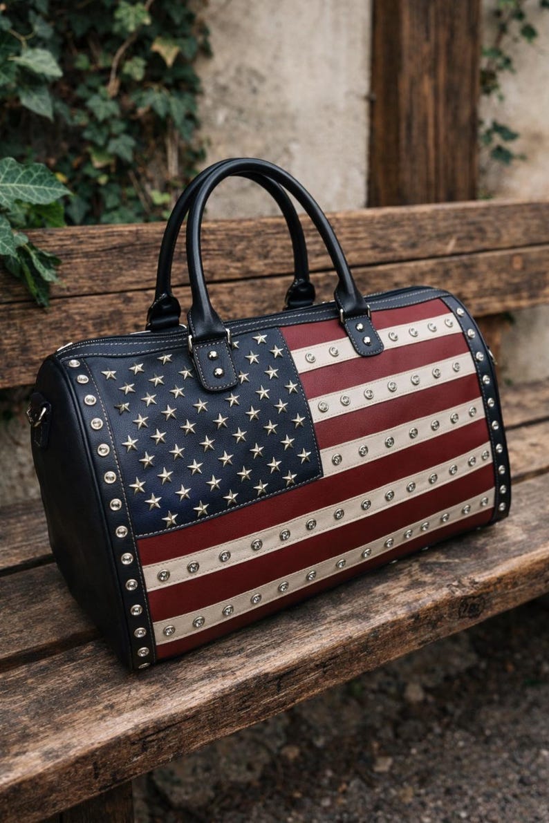 May include: A black, red, and white duffel bag with the American flag design. The bag features a navy blue section with gold stars, red and white stripes, and silver studs around the edges and on the flag design. It has black handles.
