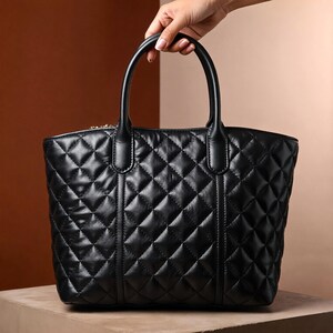 Puede incluir: Bolso tote negro acolchado con dos asas superiores. El bolso presenta un patr&oacute;n de diamantes y un cierre de cremallera. El bolso est&aacute; sostenido por una mano y se encuentra sobre un fondo neutro.