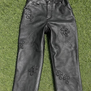 May include: Black leather trousers featuring embroidered cross motifs. The high-waisted trousers have a straight leg cut. Cross designs are stitched on the front and back, providing a distinctive look to the garment.