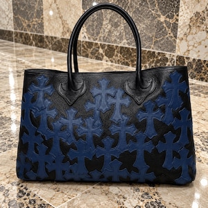 Puede incluir: Un bolso tote de cuero negro con numerosos parches en forma de cruz azul cosidos en la superficie frontal. El bolso incluye dos asas de cuero negro resistentes unidas a la parte superior, con un acabado granulado en la base.