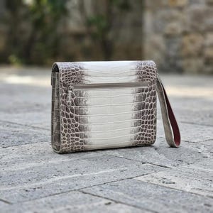 Puede incluir: Un bolso de mano rectangular con un patrón texturizado en relieve de piel de cocodrilo en tonos blanco y marrón oscuro. El bolso incluye un bolsillo horizontal con cremallera en el exterior y una correa de muñeca a juego con forro rojo oscuro.