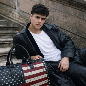 May include: A black leather duffel bag with the American flag design. The bag features a navy blue field of stars and red and white stripes, accented with silver studs. The bag has black handles and trim.