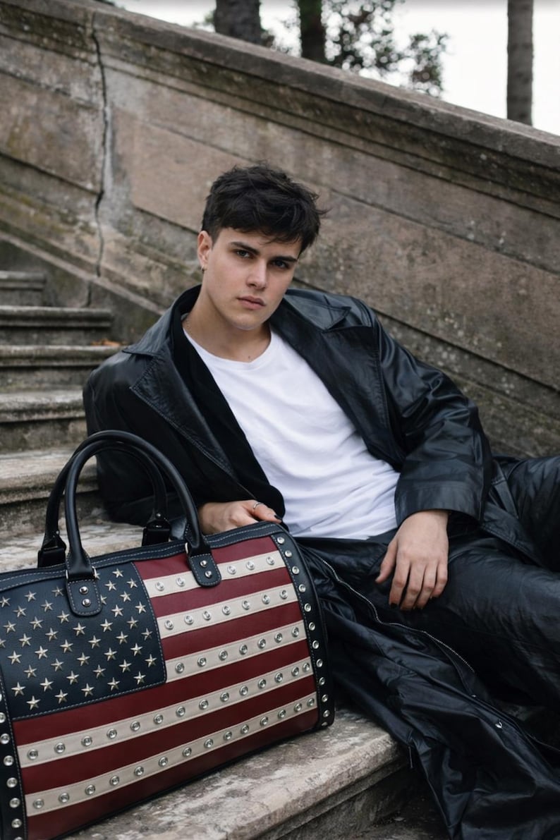 May include: A black leather duffel bag with the American flag design. The bag features a navy blue field of stars and red and white stripes, accented with silver studs. The bag has black handles and trim.
