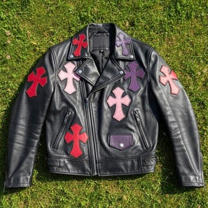 Może przedstawiać: Czarna skórzana kurtka z naszywkami w kształcie czerwonych, różowych i fioletowych krzyży. Kurtka ma zapięcie na zamek błyskawiczny, małą kieszeń i klasyczny krój motocyklowy. Leży na zielonej trawie.