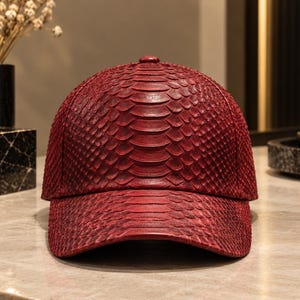 Puede incluir: Una gorra de béisbol de color rojo intenso con un patrón texturizado similar a escamas en toda su superficie. La gorra presenta una corona estructurada y una visera curva, con un diseño consistente y repetitivo que imita las escamas.