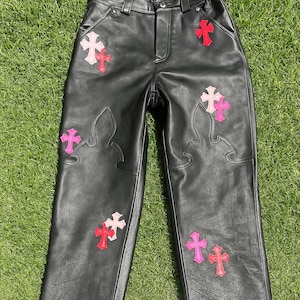 May include: Black leather trousers featuring pink and red cross embellishments. The trousers have a classic five-pocket design and a straight leg. The crosses are in different sizes and are placed on the front and back of the trousers.