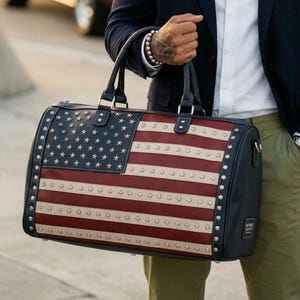 May include: A duffel bag featuring the American flag design. The bag has a navy blue body with red and white stripes and a field of white stars. Silver studs line the edges. Black handles and accents complete the design.