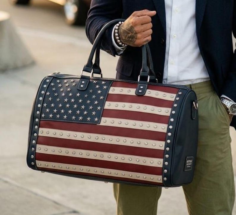 May include: A duffel bag featuring the American flag design. The bag has a navy blue body with red and white stripes and a field of white stars. Silver studs line the edges. Black handles and accents complete the design.