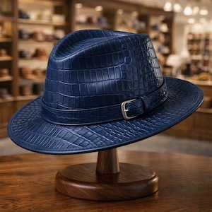 Puede incluir: Un sombrero fedora azul marino con un patr&oacute;n texturizado similar al de un cocodrilo. El sombrero presenta una banda a juego con una hebilla plateada. Se exhibe en un soporte de madera en una tienda de sombreros.