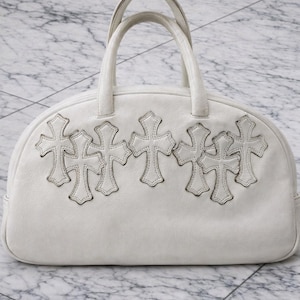 Puede incluir: Un bolso de mano de cuero blanco con una silueta redondeada en la parte superior y dos asas. La parte frontal del bolso está decorada con cinco aplicaciones cosidas en forma de cruz dispuestas en una fila horizontal en la sección superior.