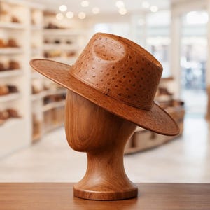 May include: A brown leather hat with a textured pattern, displayed on a wooden mannequin head. The hat has a wide brim and a structured crown. The background shows a shop with shelves of hats.
