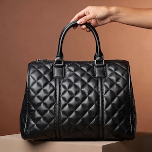 Puede incluir: Bolso negro acolchado con asa superior y cierre de cremallera. El bolso presenta un patr&oacute;n de diamantes y herrajes dorados. El bolso est&aacute; sostenido por una mano.