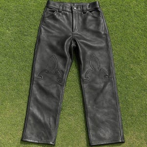 Puede incluir: Pantalones de cuero negro con costuras decorativas en la parte delantera. Los pantalones tienen un diseño clásico de cinco bolsillos y cierre de botón y cremallera. Los pantalones se muestran sobre una superficie de hierba verde.
