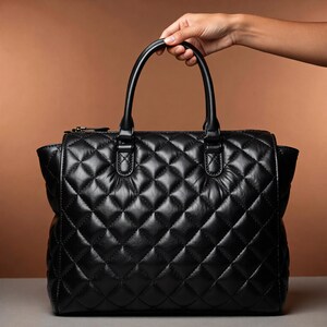 Puede incluir: Bolso negro acolchado con un patr&oacute;n de diamantes. El bolso tiene dos asas cortas y un cierre de cremallera. El bolso est&aacute; sostenido por una mano. El fondo es de color marr&oacute;n liso.