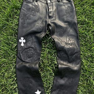 May include: Black leather pants featuring a button closure, belt loops, and multiple cross-shaped patches stitched across the front. The design includes a single white cross patch on the right thigh and a white star patch on the lower left leg.