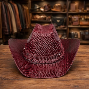 Op de afbeelding: Een diepbordeauxrode cowboyhoed met een gestructureerd slangenleerpatroon over het gehele oppervlak. De hoed is voorzien van een gevlochten band rond de basis van de kroon, geaccentueerd met kleine zilverkleurige metalen studs langs de onderrand.
