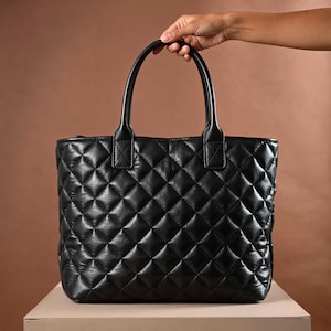 Puede incluir: Bolso tote negro acolchado con un patr&oacute;n de diamantes. El bolso tiene dos asas y un cierre de cremallera. El bolso est&aacute; hecho de un material suave similar al cuero. El bolso est&aacute; sobre una superficie beige.