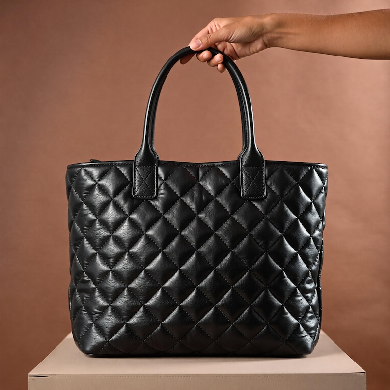 Puede incluir: Bolso tote negro acolchado con un patr&oacute;n de diamantes. El bolso tiene dos asas y un cierre de cremallera. El bolso est&aacute; hecho de un material suave similar al cuero. El bolso est&aacute; sobre una superficie beige.