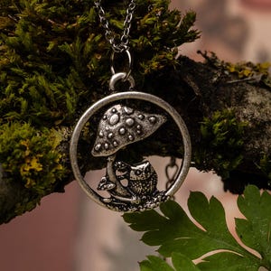 May include: A silver-toned pendant necklace featuring a mushroom and owl design within a circular frame. The pendant hangs from a delicate chain, displayed on a moss-covered branch with green foliage. The necklace is a nature-themed accessory.