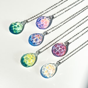 Puede incluir: Seis collares con colgantes de cristal de colores y cadenas plateadas. Cada colgante, con forma de lágrima, muestra un mosaico de colores como verde, azul, morado y amarillo. Los collares se exhiben sobre una superficie blanca.