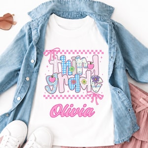 May include: A white t-shirt featuring the text "third grade" in a blue and pink checkered pattern with small flower and apple icons, the name "Olivia" in pink script, a blue denim button-down shirt, a pink tiered skirt, and heart-shaped sunglasses.