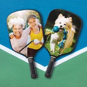 May include: Two pickleball paddles with black handles. One paddle features two smiling girls, the other shows a dog with a blue and yellow toy. The paddles are on a blue and green surface with white lines.