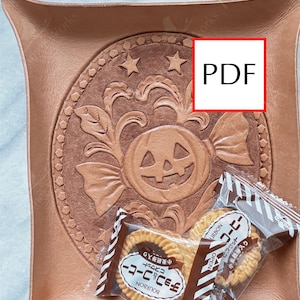 Peut inclure: Un vide-poche carré en cuir beige aux coins repliés, orné d'un motif central gaufré représentant une citrouille d'Halloween entourée de feuilles et d'étoiles. Un emballage plastique transparent contenant deux biscuits ronds Bourbon repose dessus.