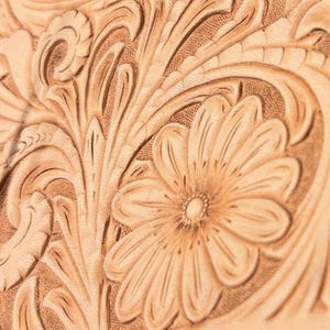 May include: Close-up of intricately carved leather, showcasing a floral design with a prominent daisy. The natural tan leather is detailed with embossed patterns of leaves and petals, creating a textured, three-dimensional effect. The craftsmanship highlights a rustic, handcrafted aesthetic.