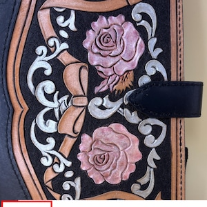 Könnte beinhalten: Schwarzer Leder-Journal-Einband mit geprägten Rosen, Bändern und Schnörkeln. Das Design zeigt rosa Rosen, hellbraune Bänder und silberne Schnörkel auf schwarzem Hintergrund. Das Journal hat einen schwarzen Lederriemenverschluss. Der Text "Leather Works THALIA" ist sichtbar.