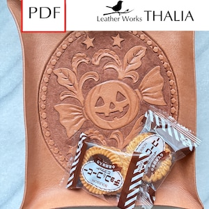 May include: A square, light brown leather tray with a carved Halloween design featuring a pumpkin and stars. The tray holds two packages of Japanese coffee biscuits. The text "Leather Works THALIA" is visible.