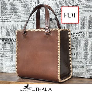 May include: A structured brown leather tote bag featuring a rectangular silhouette, two top handles with adjustable silver-tone buckles, and light-colored whipstitched edges along the side panels. The bag has a smooth finish and clean lines.