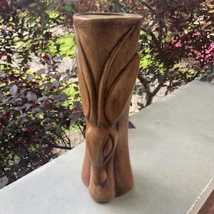 May include: A tall, brown, carved wooden vase with a botanical design. The vase features a flared top and a base with three rounded feet. The carved design includes long, curved leaves and a central, bulbous shape, creating a natural, organic aesthetic.