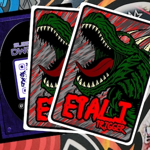 May include: Two trading cards with a green monster illustration, red tongue, and the text "ETAL J TRIGGER". A purple card sleeve with a QR code is visible. The cards have a white border and are on a colorful background.