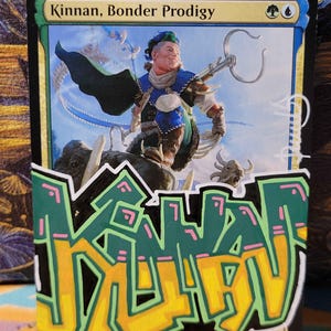 Puede incluir: Una carta coleccionable de Magic: The Gathering titulada Kinnan, Bonder Prodigy que muestra a un personaje montando una bestia, con un gran graffiti pintado a mano en verde, amarillo y rosa sobre la mitad inferior de la carta.