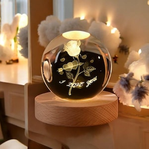 May include: A clear glass sphere with a glowing rose design and the words "I love you" etched on the surface. The sphere sits on a round wooden base, illuminated from within. The overall effect is a romantic, decorative piece.