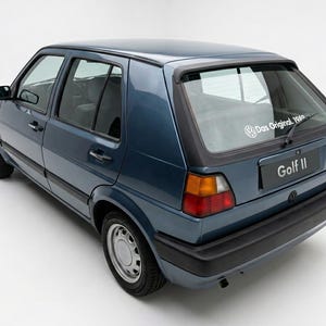 Könnte beinhalten: Ein blaues Volkswagen Golf II Schrägheckauto. Das Auto hat eine schwarze Heckstoßstange, schwarze Zierleisten und graue Räder. Die Heckscheibe hat einen weißen Aufkleber mit dem Text "Das Original 1980".