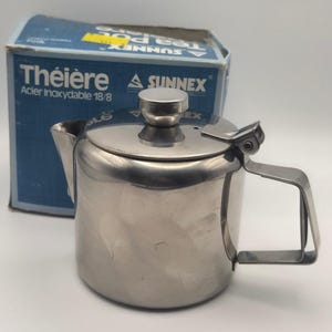 May include: A stainless steel teapot with a hinged lid, a cylindrical body, and a side handle. The teapot is positioned in front of its original blue and white cardboard box, which features the text Sunnex, Théière, and Acier Inoxydable 18/8.