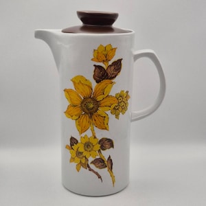 May include: A white ceramic pitcher with a brown lid and a handle. The body of the pitcher features a floral pattern with yellow sunflowers and brown leaves. The design is vertically oriented, showcasing a large central flower with smaller blooms.