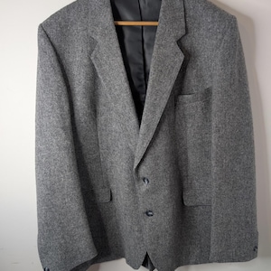 May include: A grey wool blazer featuring a textured herringbone pattern, a notched lapel, two front buttons, two flap pockets, and a chest pocket. The interior is lined with black fabric and includes a label that reads Made in England.