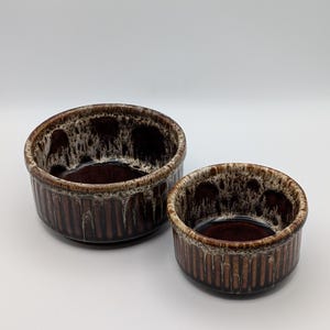 May include: Two round ceramic bowls of different sizes featuring a dark brown base colour with vertical ribbed textures on the exterior and a decorative cream and brown drip glaze pattern around the upper rim and interior edges.