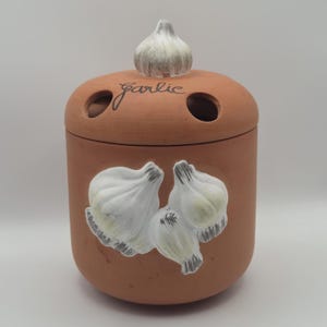 May include: A terracotta garlic keeper with a lid and a garlic-shaped knob. The container features a painted garlic bulb design and the word "Garlic" in script. The keeper has two small holes on the top for ventilation.