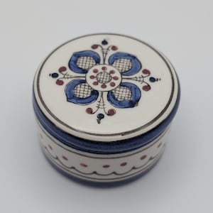 May include: A round ceramic trinket box with a lid. The lid is white with a blue floral design and red accents. The box has a blue band around the middle and a white base with red dots. The box is approximately 7.6 cm in diameter.