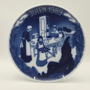Peut inclure: Une assiette décorative ronde en porcelaine blanche avec une illustration bleue représentant une scène de cuisine où deux personnes cuisinent, accompagnées d'une grande silhouette à chapeau pointu. Le texte JULEN 1989 est inscrit en haut.