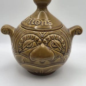 May include: A brown glazed ceramic onion storage jar with a lid featuring a knob handle. The jar has two side handles and a moulded face design with eyes, a nose, and teardrops. The word "Onions" is embossed on the lid in raised lettering.