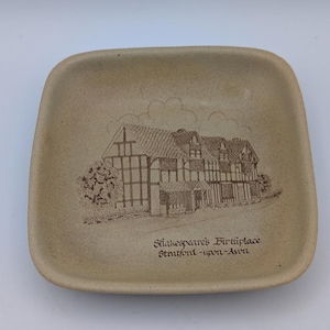 Może przedstawiać: Kwadratowa, beżowa ceramiczna miseczka z zaokrąglonymi rogami. W centrum znajduje się brązowa ilustracja liniowa budynku z muru pruskiego z napisem "Shakespeare's Birthplace Stratford-upon-Avon" wydrukowanym pod obrazkiem.